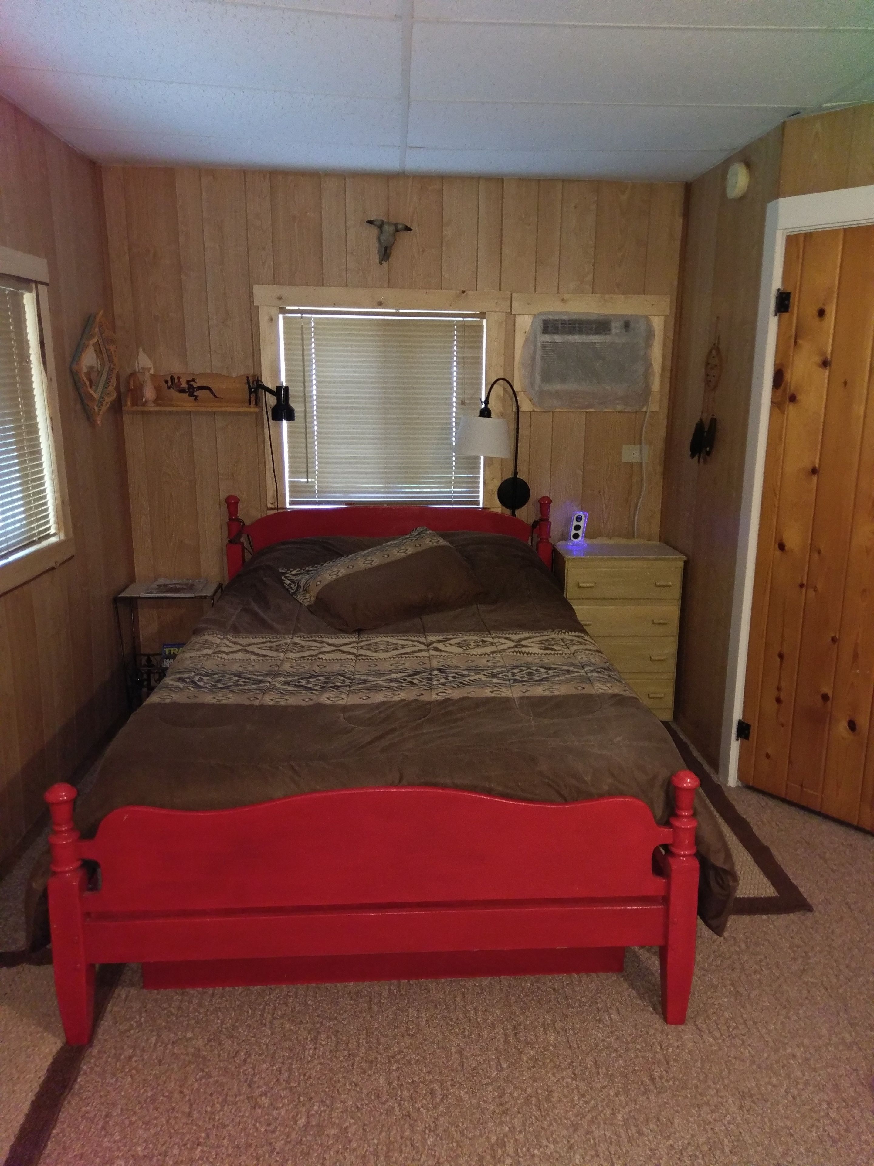 A bedroom with a red bed, wooden walls, a window with blinds, a bedside table with a lamp, and a dresser.