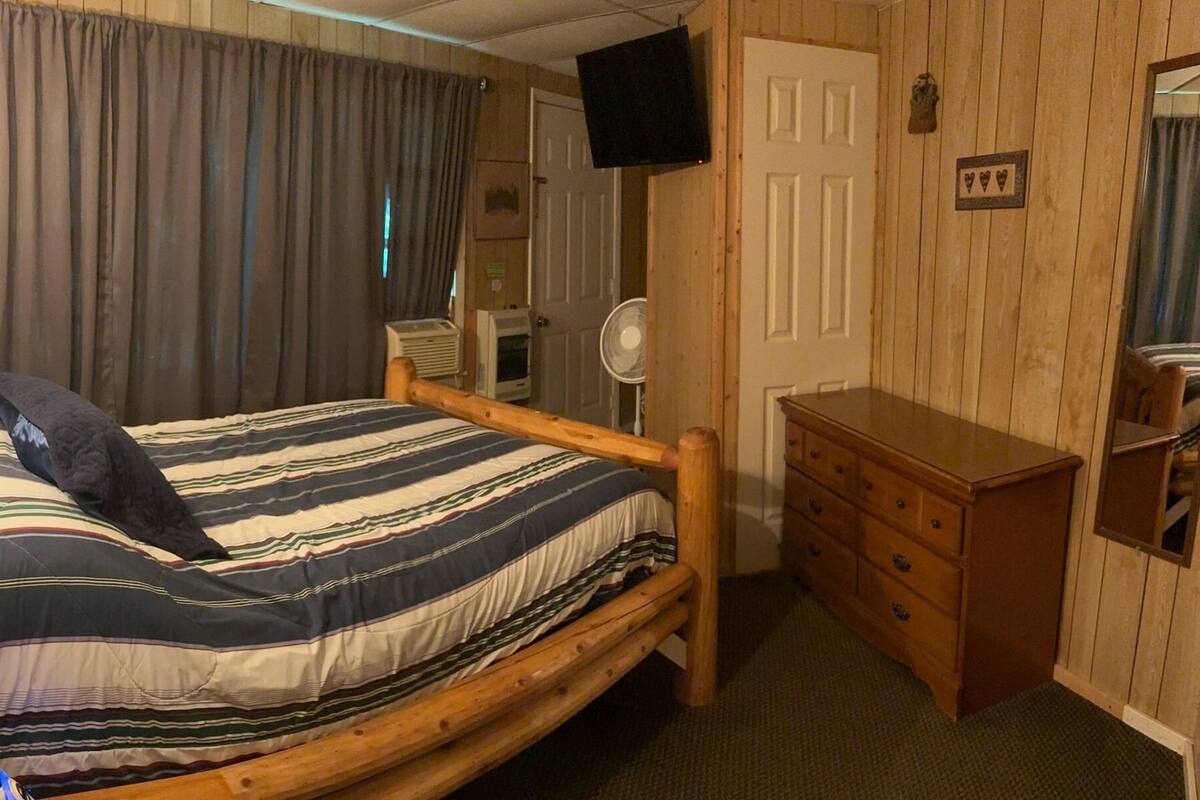 Un lit simple avec une couvre-lit à rayures, une commode en bois, un miroir, une télévision et un ventilateur dans une pièce aux murs en bois et dotée d’une porte.