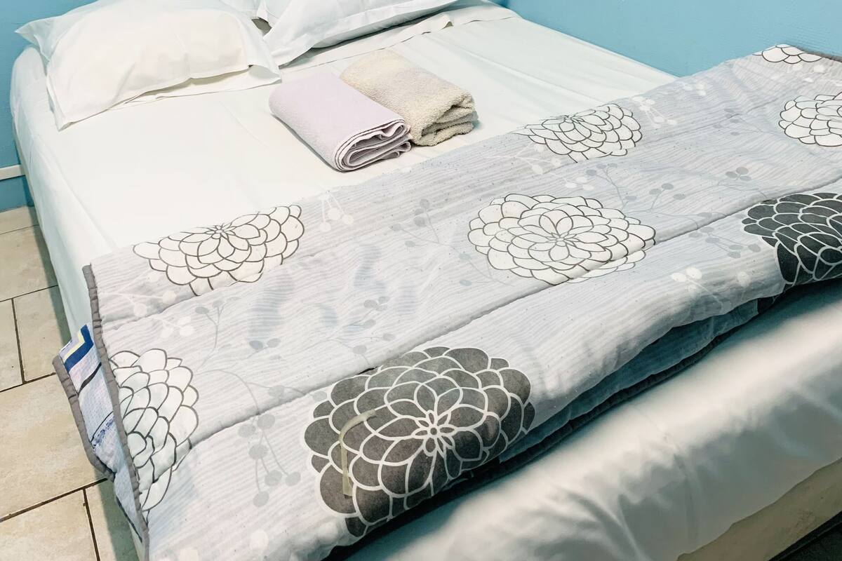 Una cama con ropa blanca y una manta de estampado floral, una toalla doblada y dos almohadas.