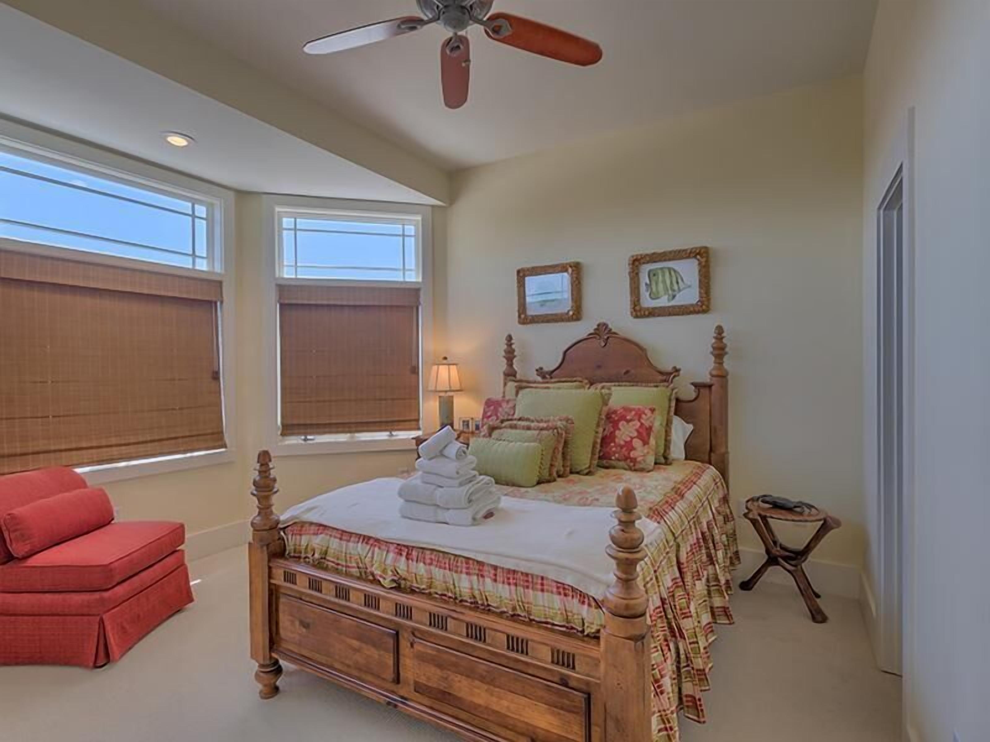 A bedroom with a wooden four-poster bed, a red chair, a small wooden stool, and two windows with blinds.