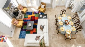 A spacious living area with a white sofa, a colorful rug, wicker chairs, a glass dining table with yellow placemats, and a decorative centerpiece.