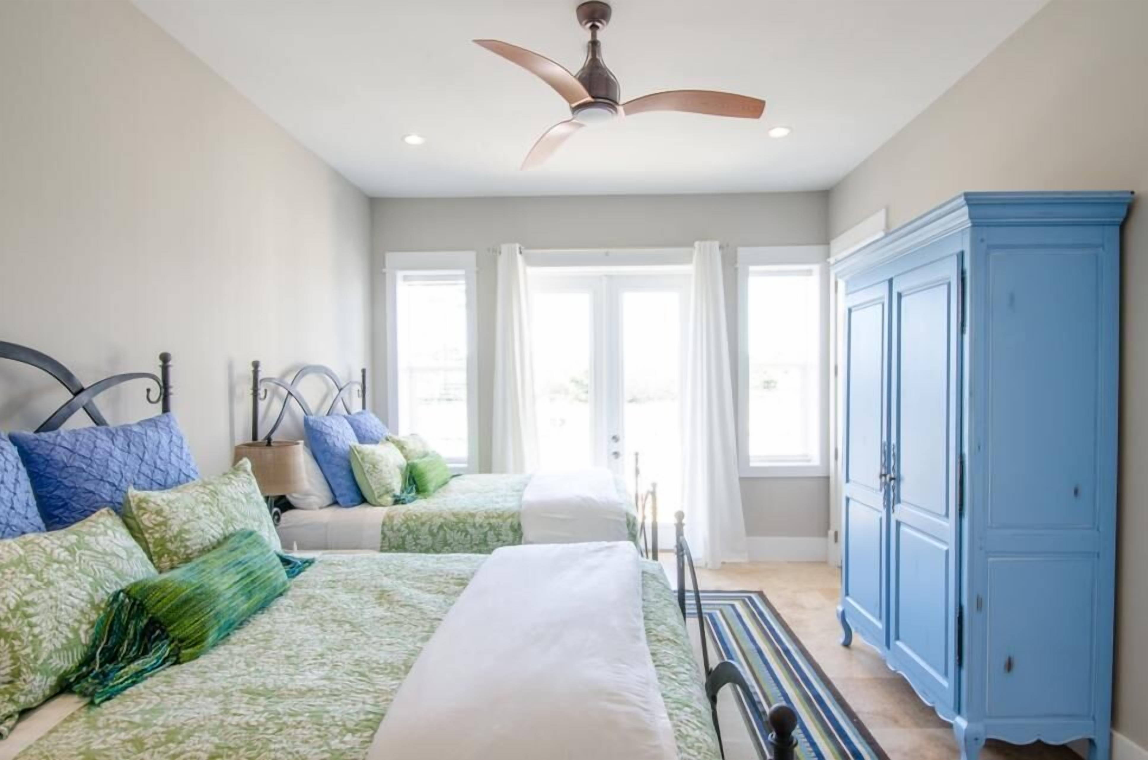 A bedroom with two beds, a ceiling fan, a blue wardrobe, and large windows.