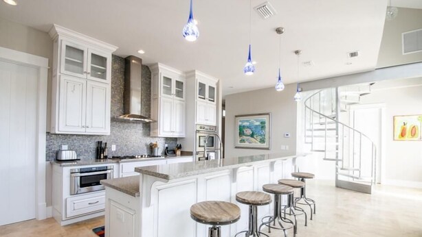 A modern kitchen with white cabinets, a large island with stools, and blue pendant lights.
