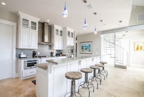 A modern kitchen with white cabinets, a large island with stools, and blue pendant lights.