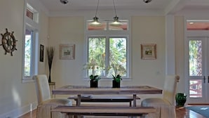 Un comedor con una mesa de madera y bancos, dos plantas en macetas y vistas a palmeras a través de la ventana.