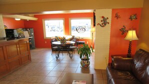 A spacious living area with a brown leather sofa, a coffee table with magazines, a dining table with chairs, a potted plant, and a kitchen area with a refrigerator and a coffee machine. - El Patio Inn (San Angelo)