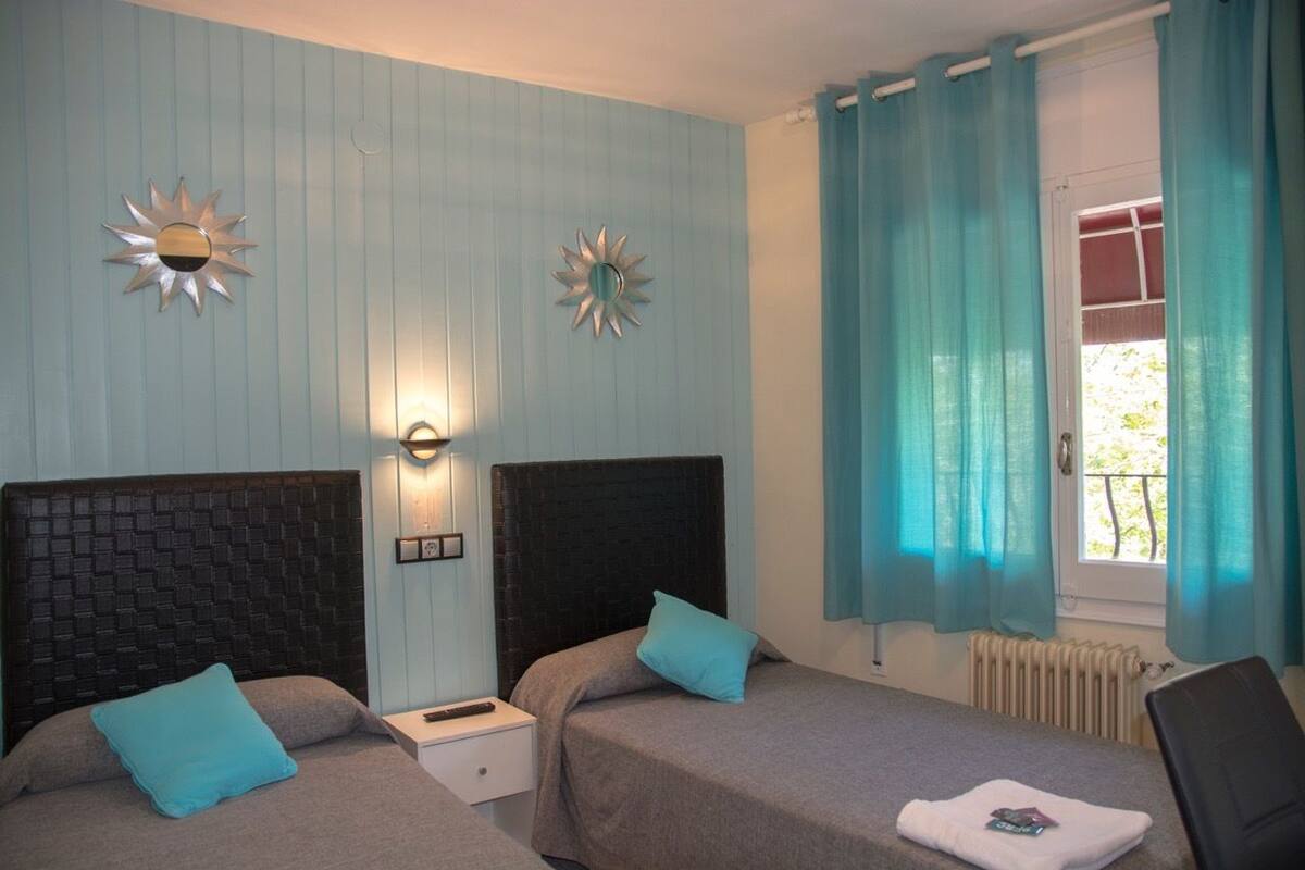 A hotel room with two beds, a nightstand, a wall-mounted light, and a window with curtains.
