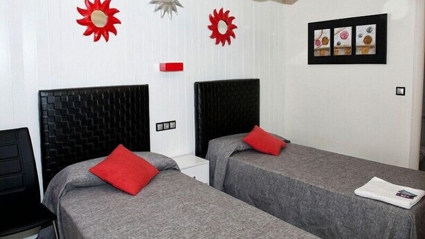 A hotel room with two beds, each with a black headboard and grey bedding, red pillows, and a white nightstand. There are decorative sun-shaped wall art pieces and a framed picture above the beds. - Hostal Emporda (Castello d'Empuries)