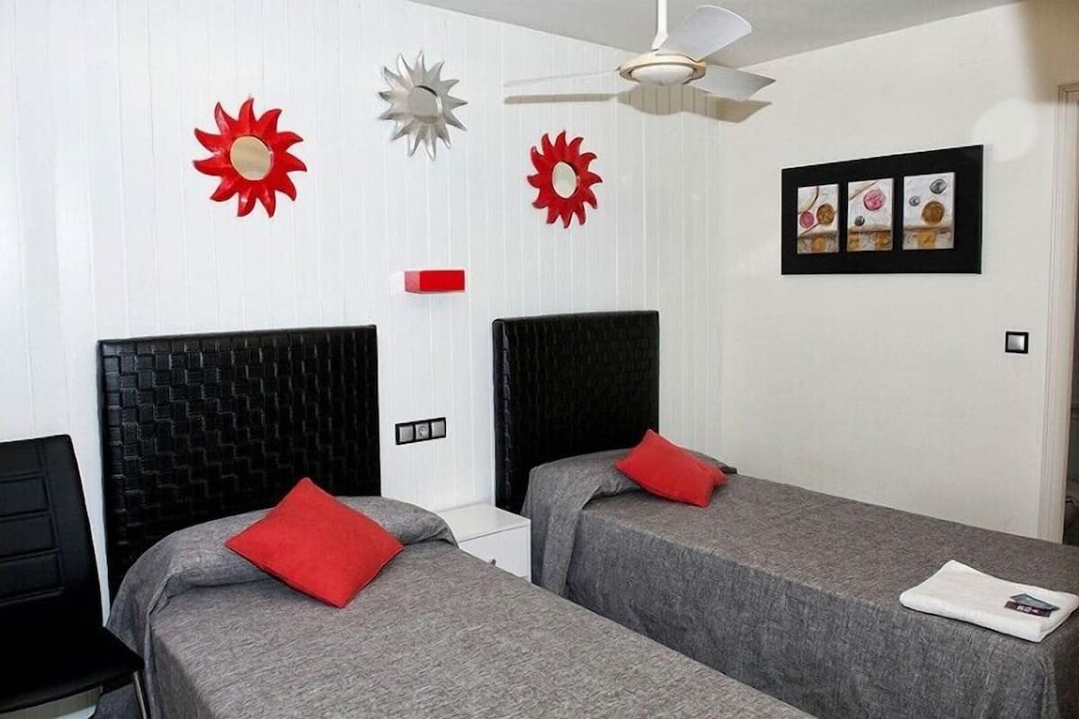 A hotel room with two beds, each with a black headboard and grey bedding, red pillows, and a white nightstand. There are decorative sun-shaped wall art pieces and a framed picture above the beds.