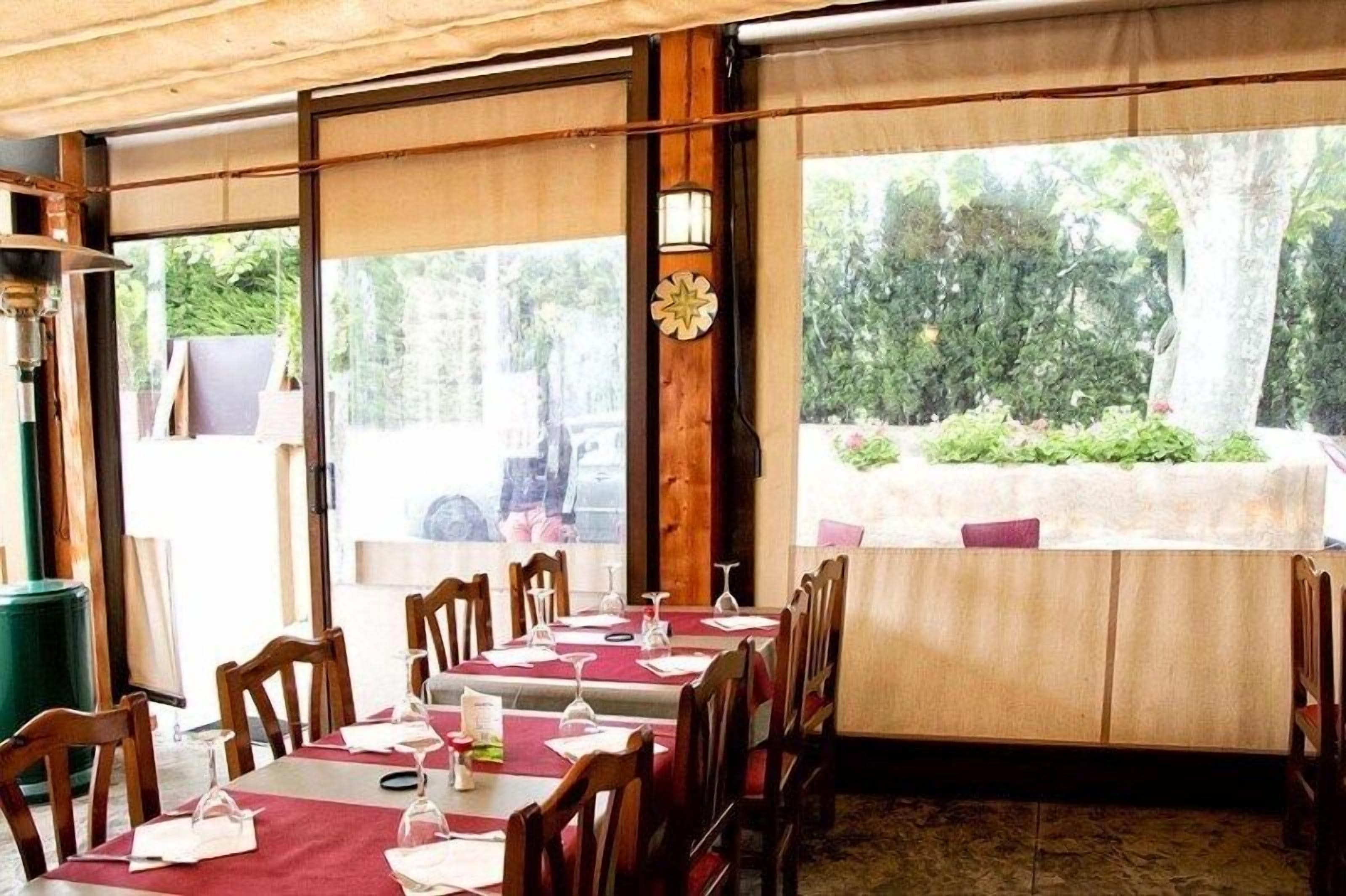 Un restaurante con mesas y sillas de madera, amplios ventanales que dan a zonas verdes, y una lámpara de pared decorativa.
