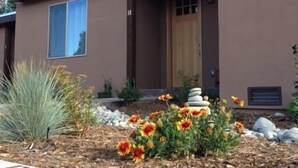 A modern two-story house with a brown exterior, a front porch, and a garden with flowers and rocks.