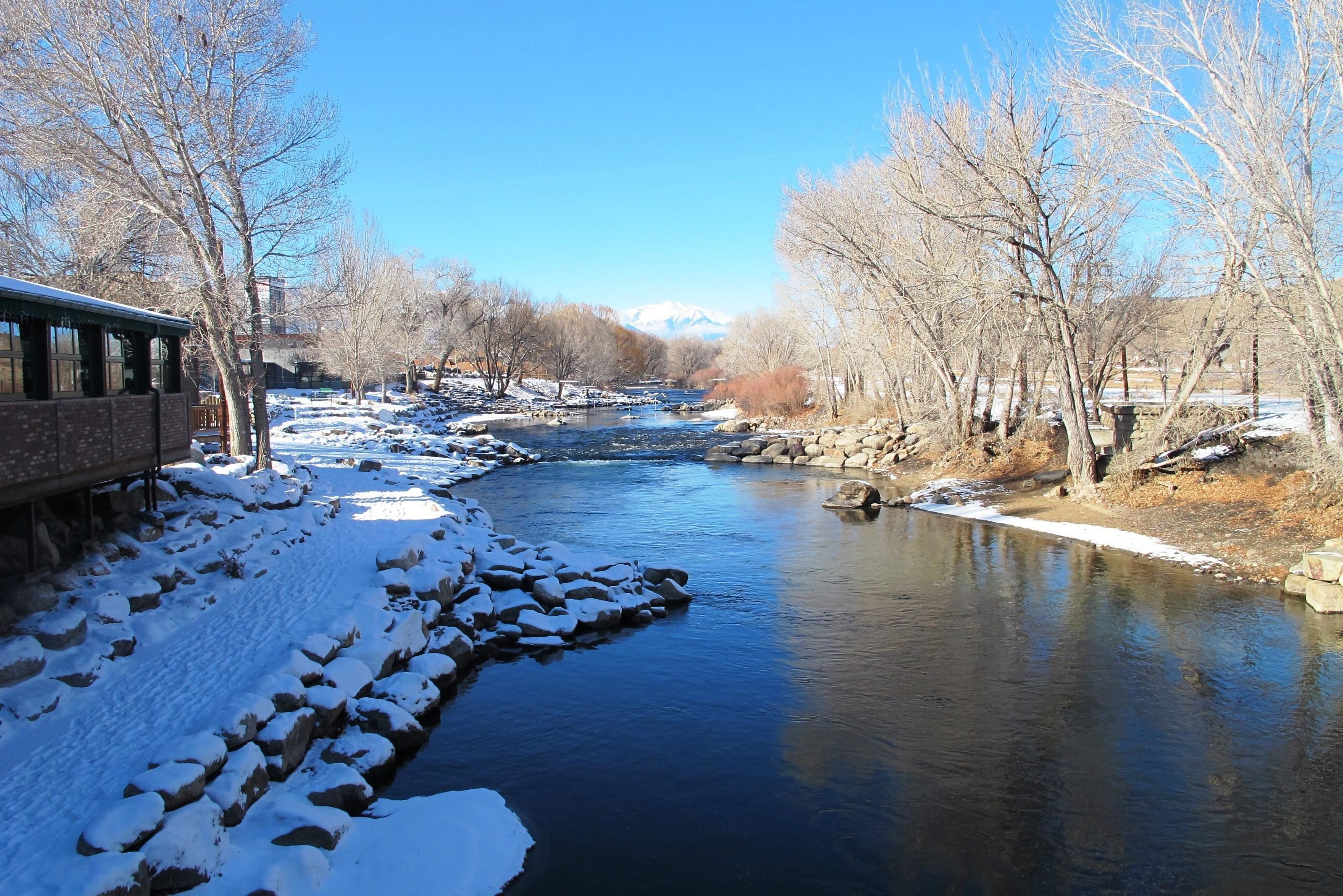 A river with snow-covered banks and trees, a building with large windows on the left, and a clear blue sky.