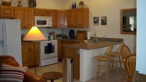 A compact kitchen with wooden cabinets, a microwave, a stove, and a small dining area with a chair and a table. - Villa on the Green 2 Bedroom Holiday Home By Pinon Vacation Rentals (Salida)