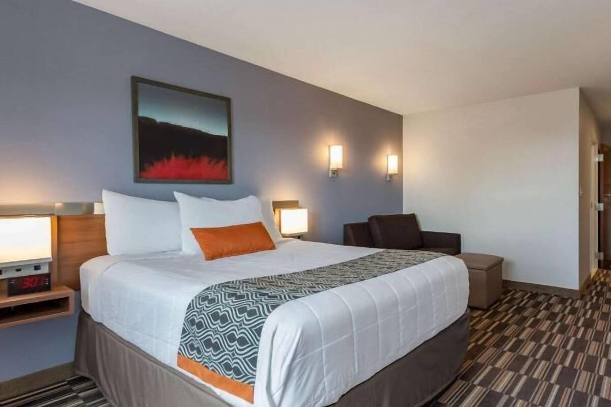 A hotel room with a bed, bedside table, lamp, wall-mounted light, and a view of the hallway.