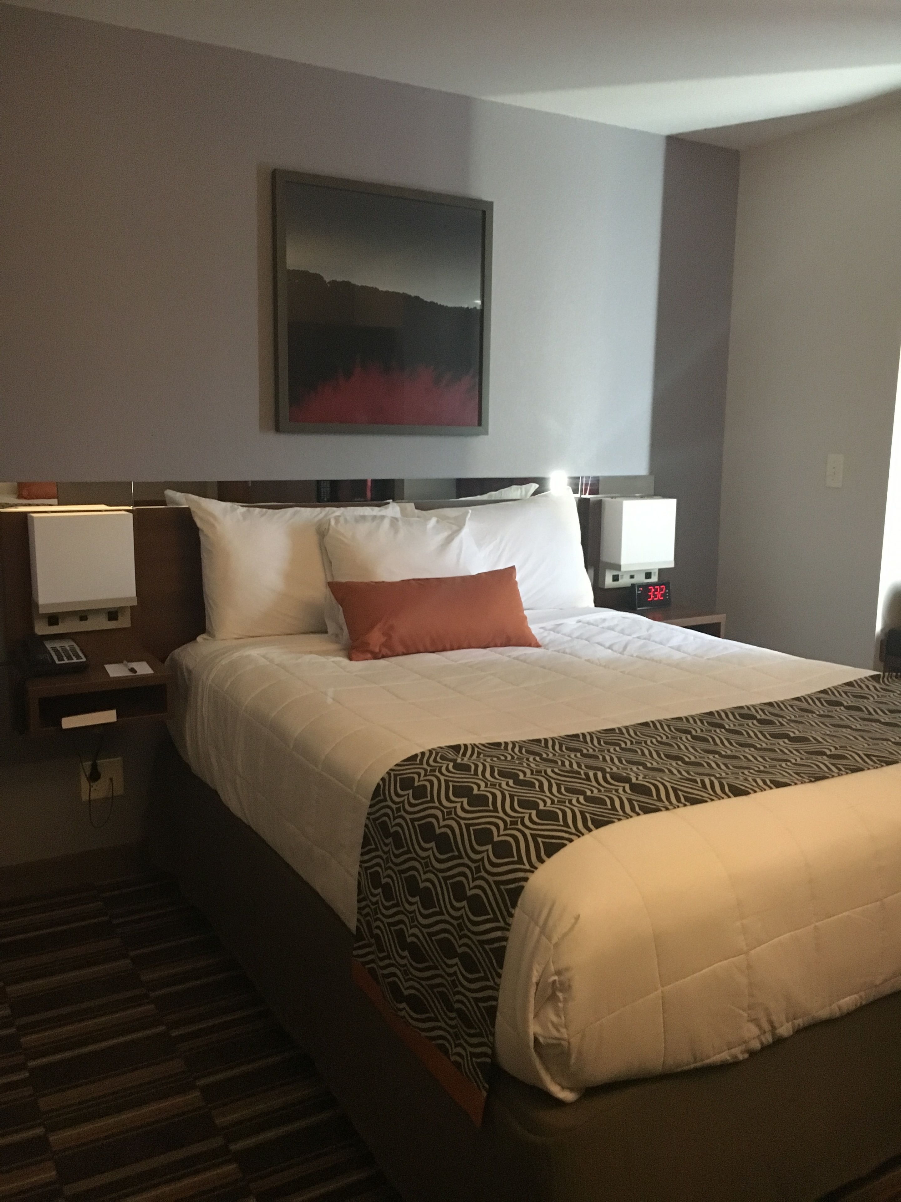 Una habitación de hotel con una cama grande, una mesita de noche con lámpara, un reloj de pared y un cuadro sobre la cama.