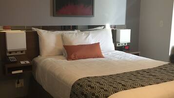 Una habitación de hotel con una cama grande, una mesita de noche con lámpara, un reloj de pared y un cuadro sobre la cama.