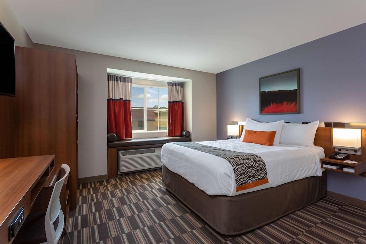 A hotel room with a bed, a desk, a chair, a window with curtains, a wall-mounted artwork, and a nightstand with a lamp.