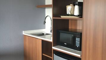 A compact kitchenette with a sink, microwave, and dishwasher, featuring wooden cabinetry and a striped carpet.