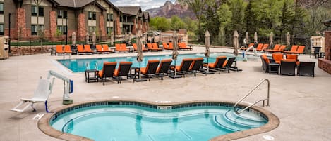 A pool area with lounge chairs and a hot tub, surrounded by a building with a view of mountains.