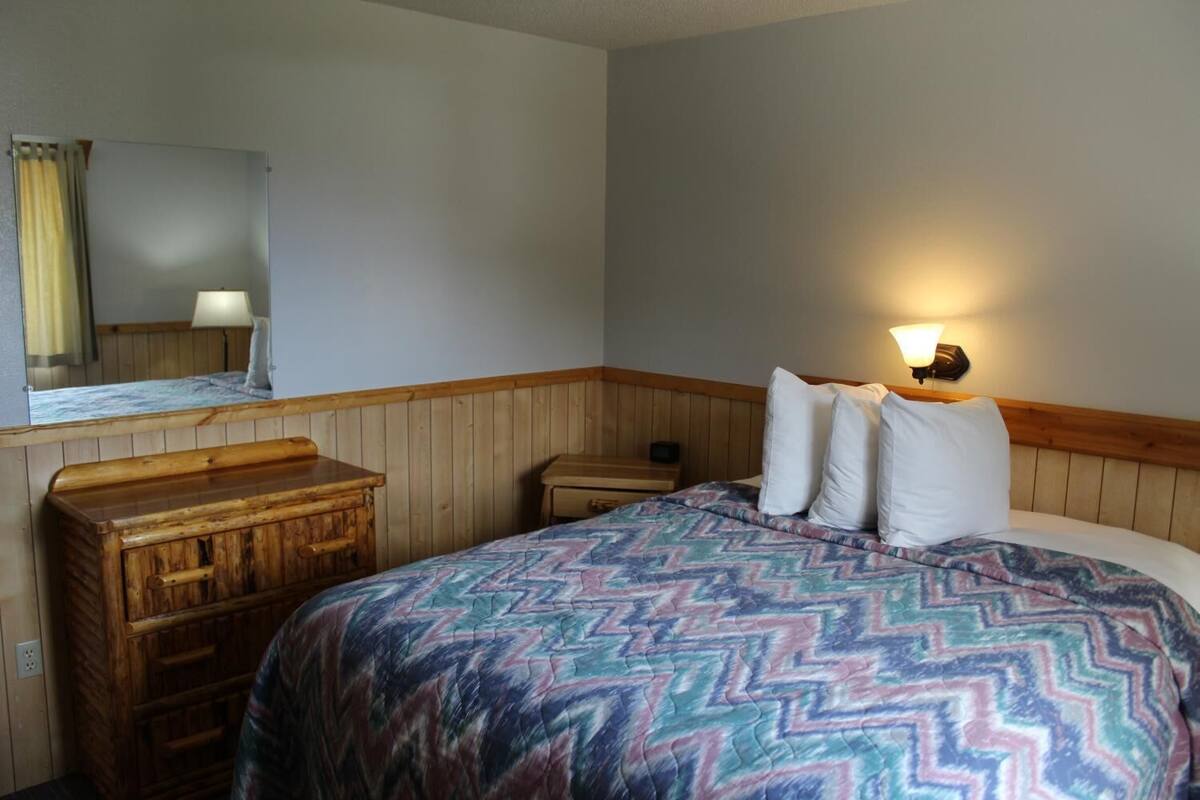 Habitación de hotel con tocador de madera, cama con manta estampada, mesita de noche, lámpara y aplique de pared.