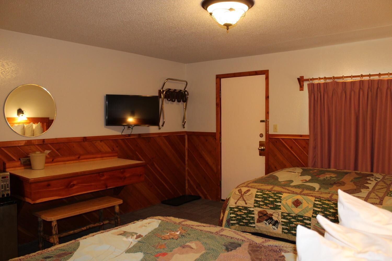 A hotel room with a wooden bench, a television, a bed with a quilt, and a door with a curtain.
