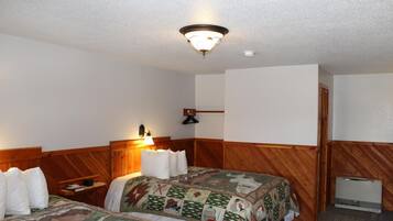 Habitación de hotel con dos camas, cabeceros de madera, una mesita de noche, una lámpara, un calentador de pared y una pared blanca lisa.