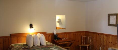 Un dormitorio con cabecera de madera, cama con colcha, escritorio con silla, luz de pared y un cuadro enmarcado.