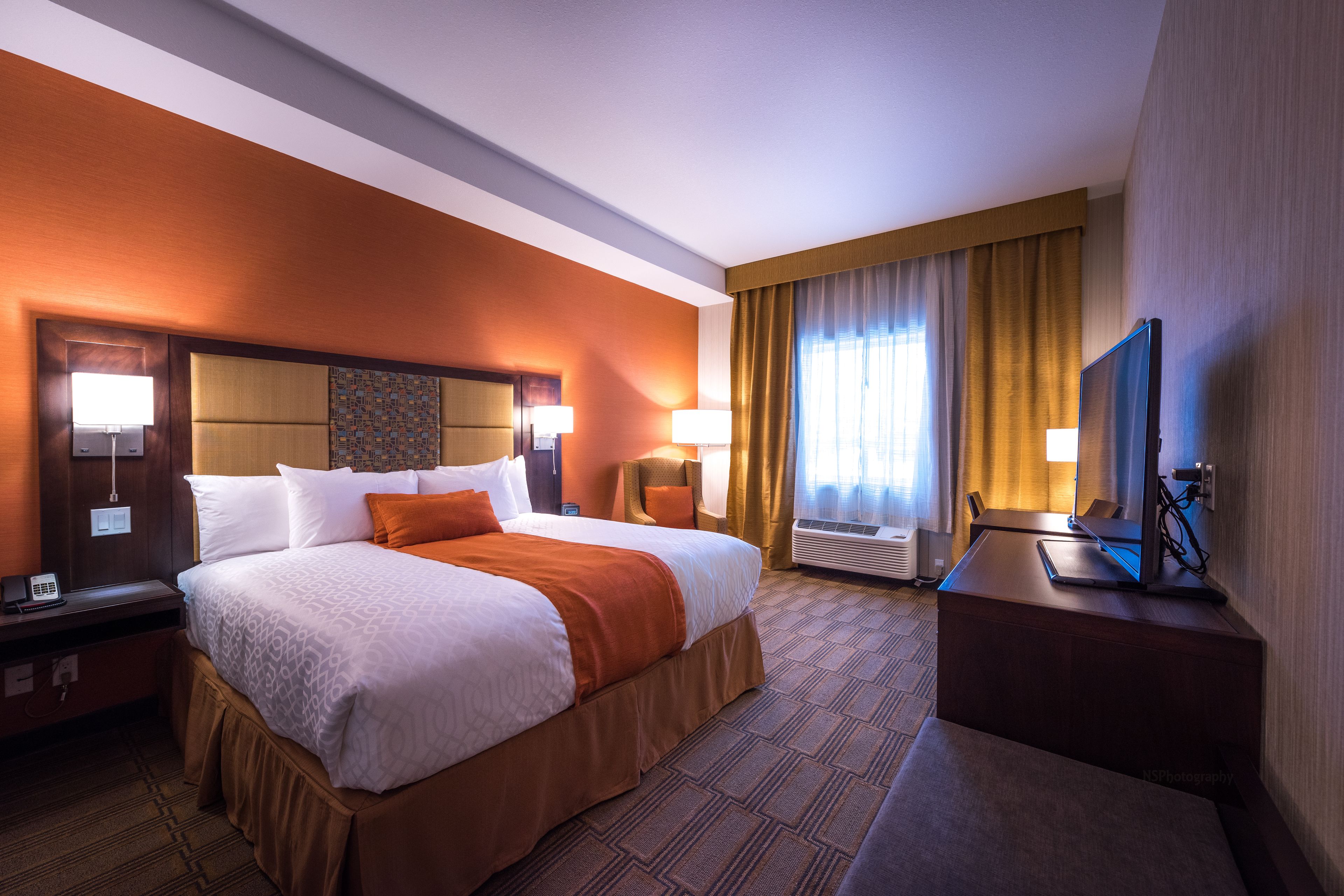 A hotel room with a large bed, two bedside tables, a desk with a chair, a flat-screen TV, and a window with curtains.
