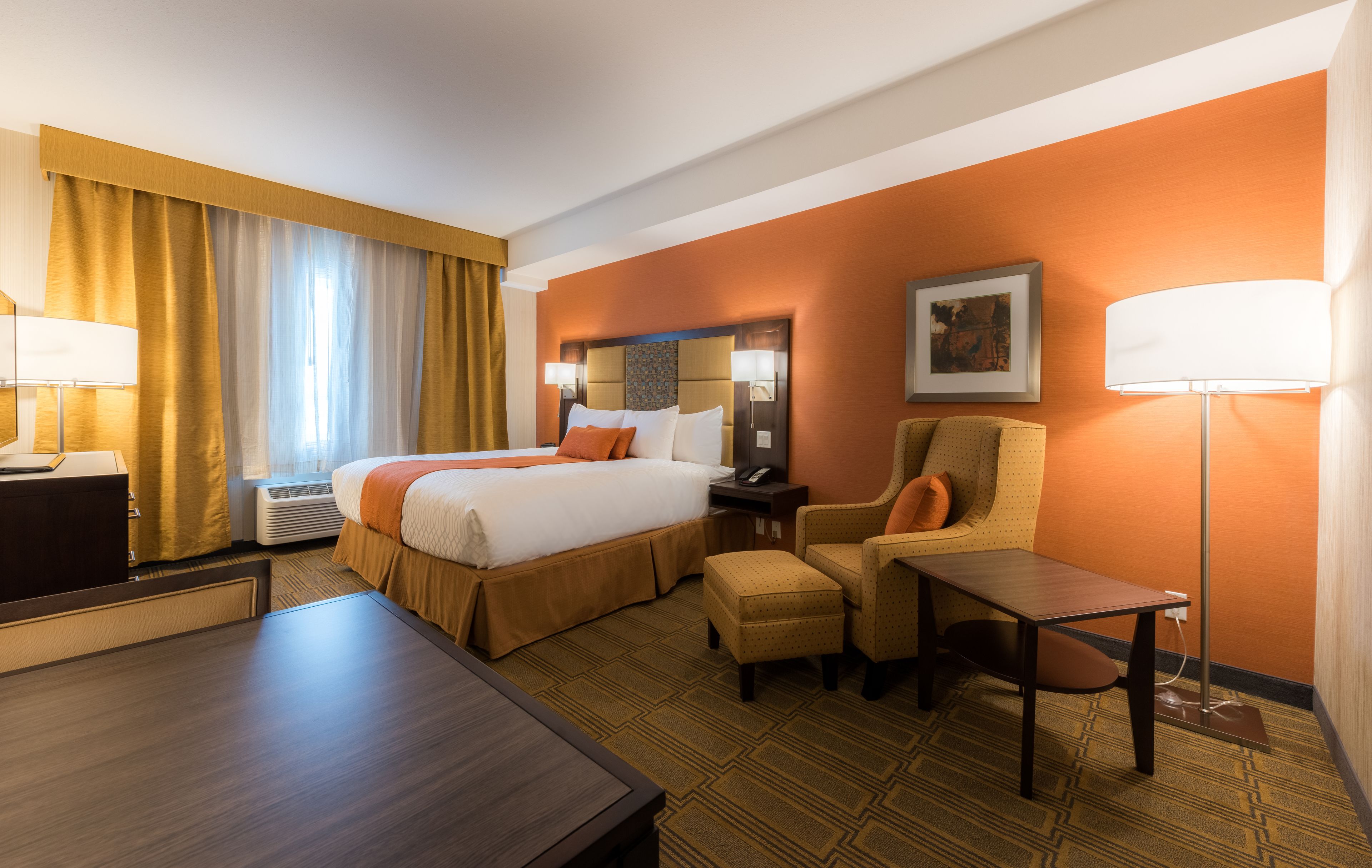 A hotel room with a large bed, a desk, a chair, and a table. There is a lamp on the desk and a picture on the wall.