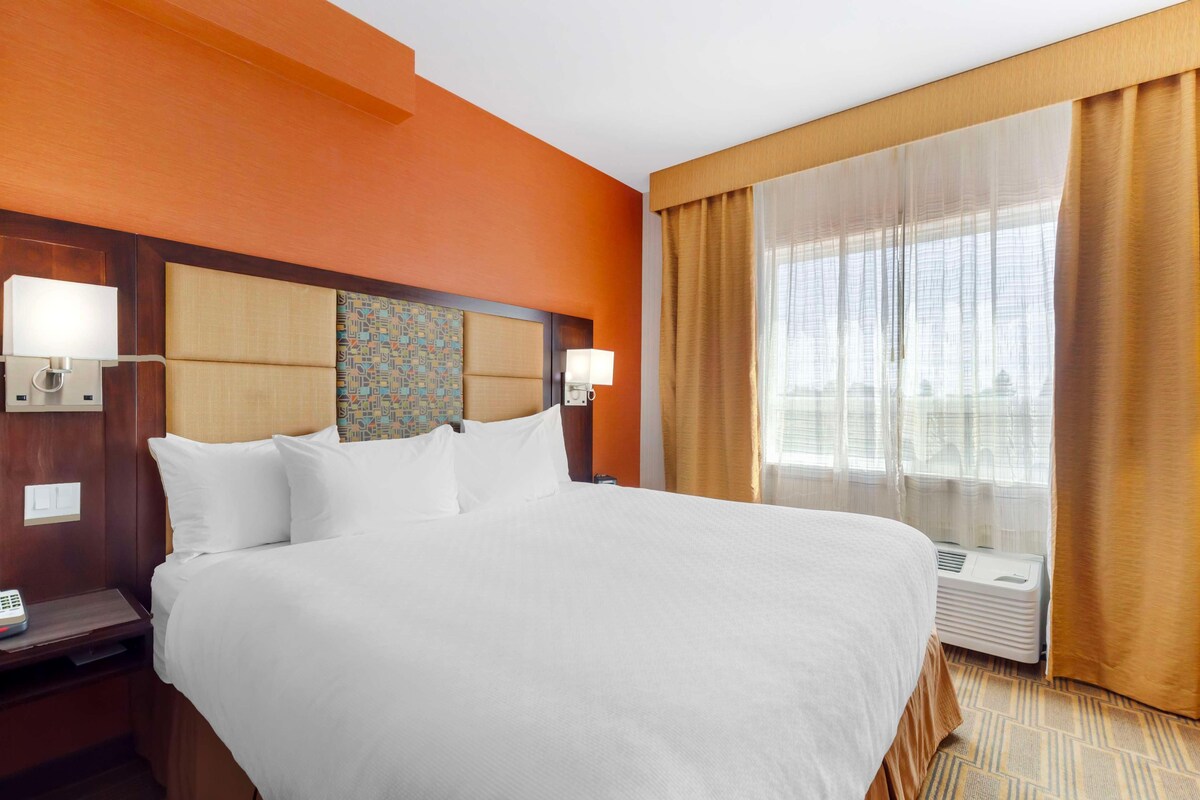 A hotel room with a large bed, two bedside tables with lamps, a headboard with a patterned design, a window with sheer curtains, and an air conditioning unit.