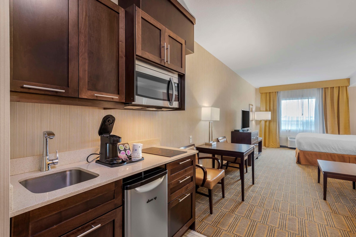 A hotel room with a kitchenette, including a microwave, coffee maker, and dishwasher. There is a bed, a desk, and a chair.