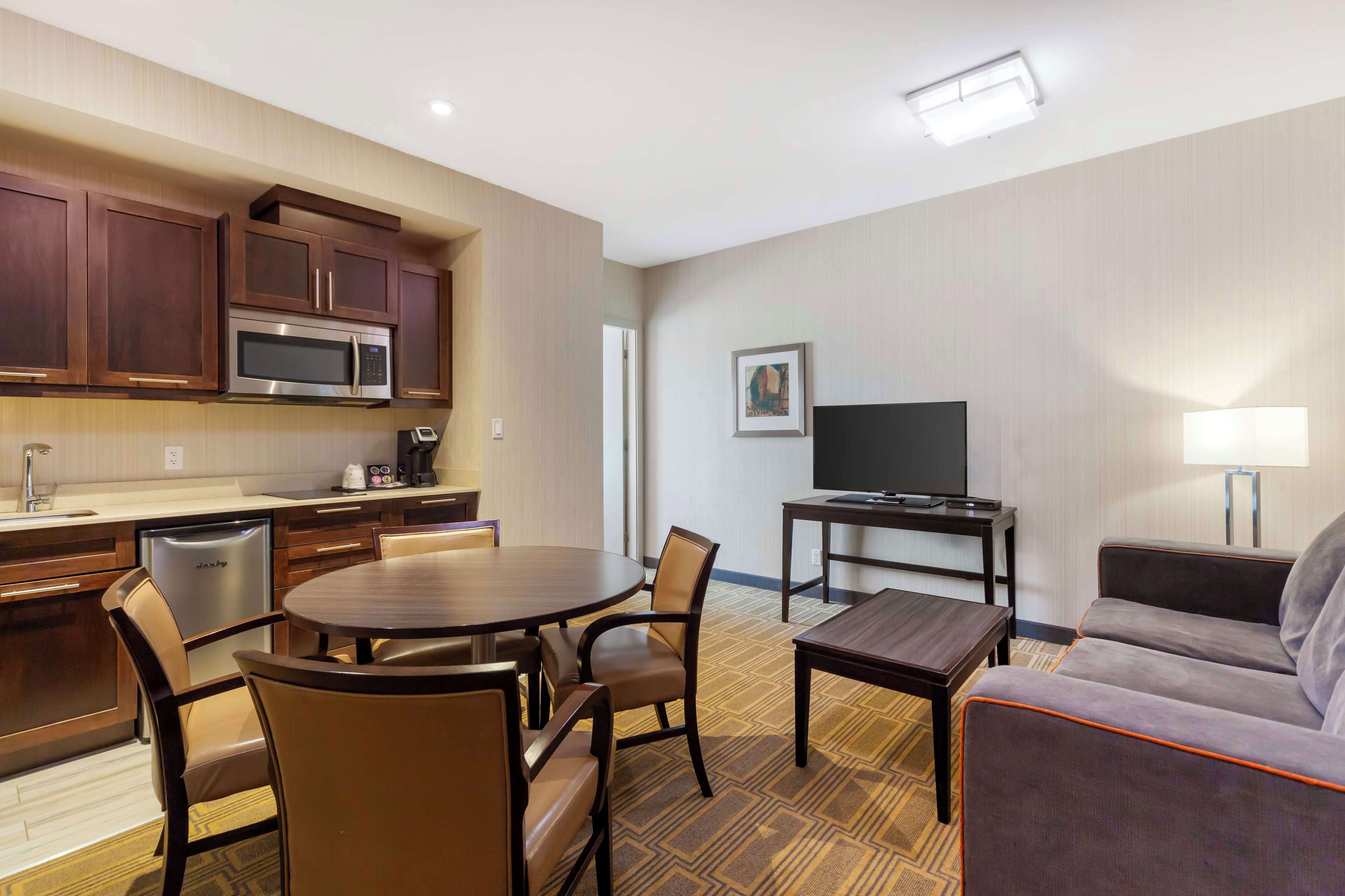 A hotel room with a kitchenette, dining area, TV, sofa, and a lamp.
