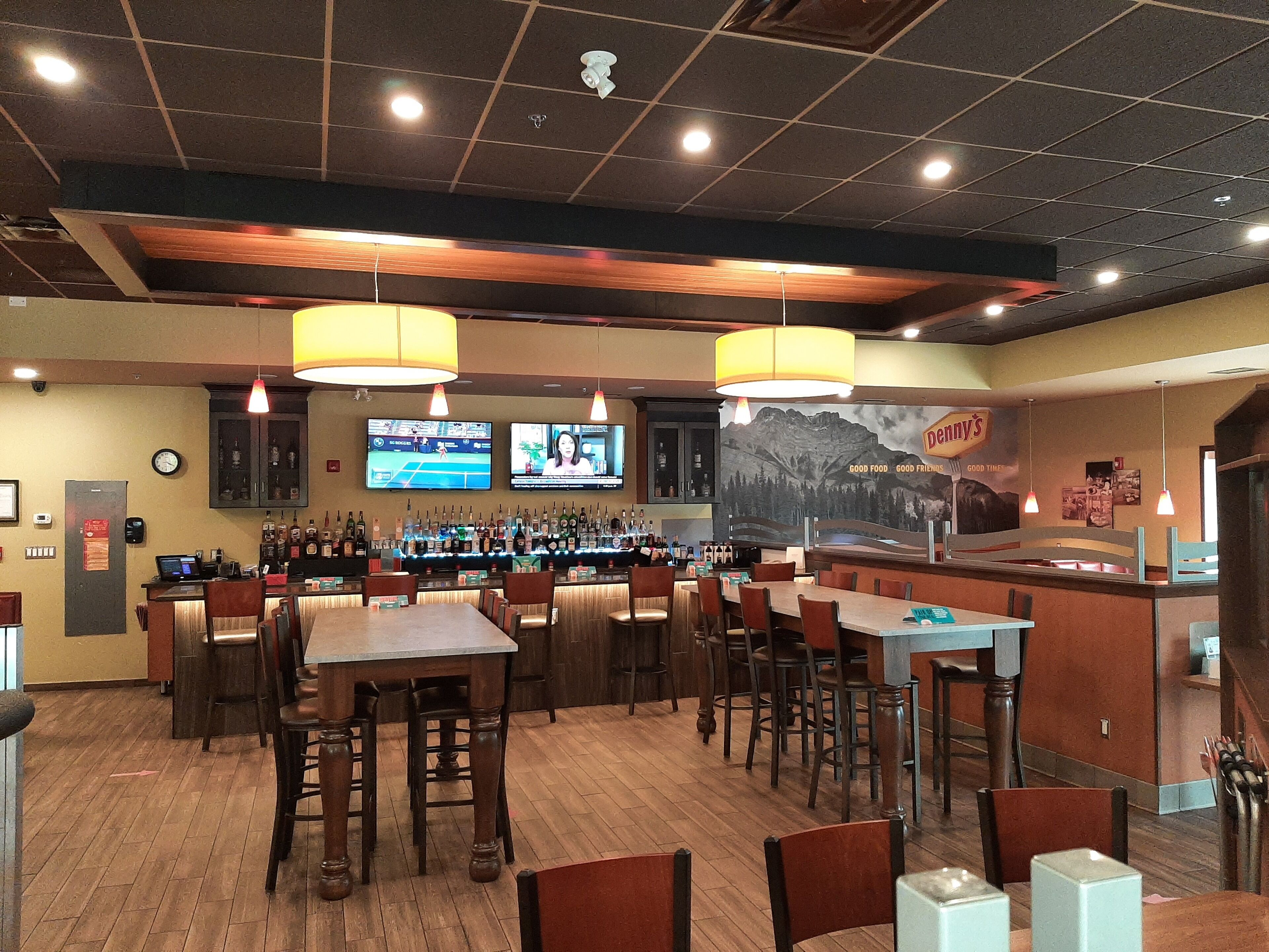 A restaurant interior with wooden tables and chairs, a bar area with bottles, and a large poster on the wall.