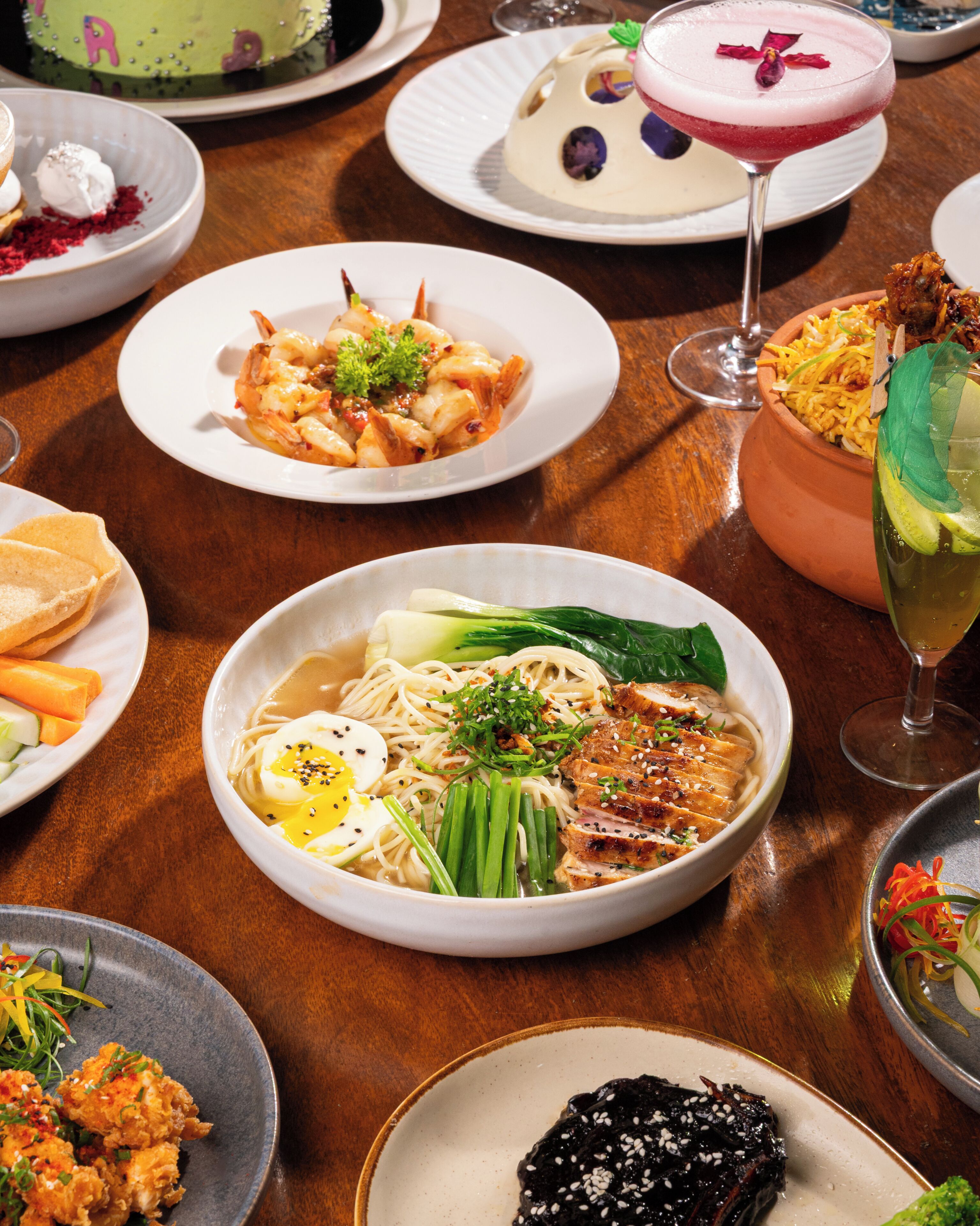 A variety of dishes on a wooden table, including a bowl of noodles with vegetables and meat, a plate of seafood, and a dessert cake.