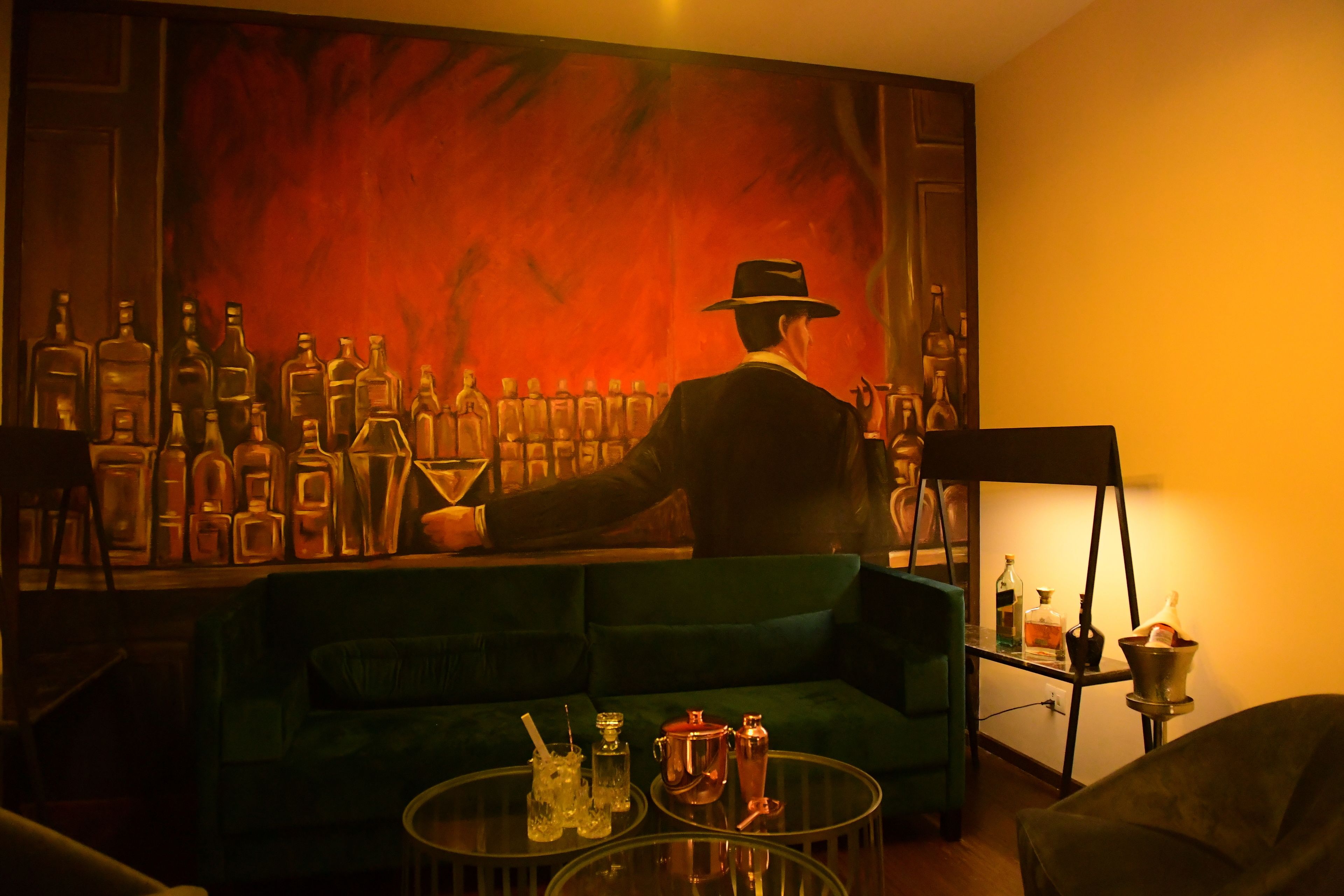 A room with a green sofa, a glass coffee table with bottles, and a painting of a man in a hat.