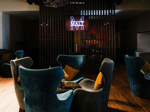 Un área de descanso moderna con sillones tapizados en azul, un panel divisor de madera con listones y un letrero de neón con las letras 'N M A L I'.