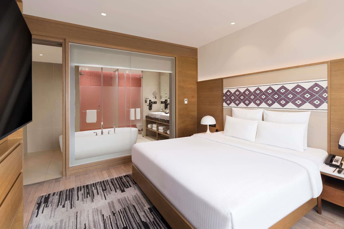 Una habitación de hotel moderna con una cama grande, un baño separado con cabina de ducha de vidrio, un cabecero de madera y una mesita de noche con lámpara.