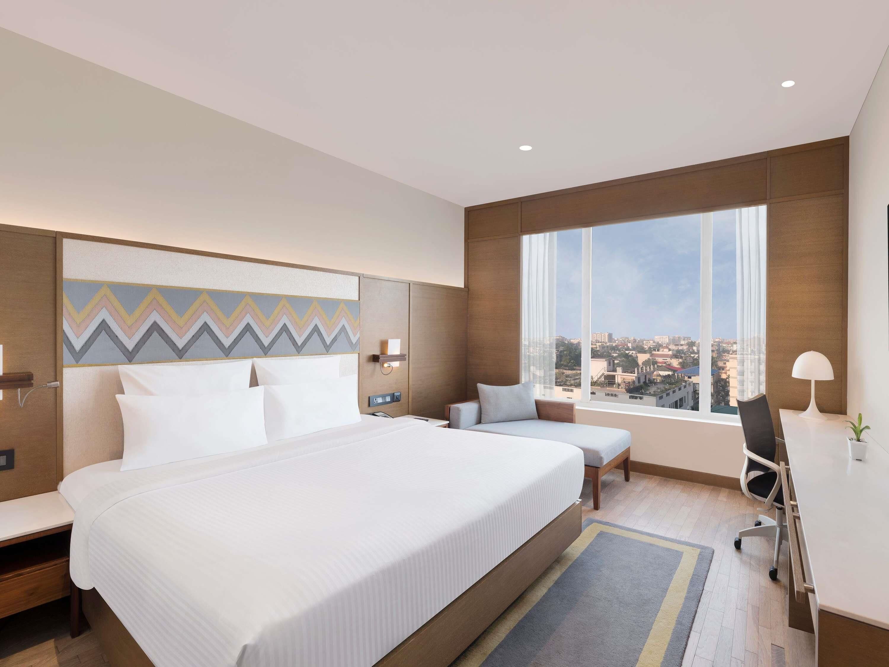 A modern hotel room with a large bed, a seating area, a desk, and a large window offering a city view.
