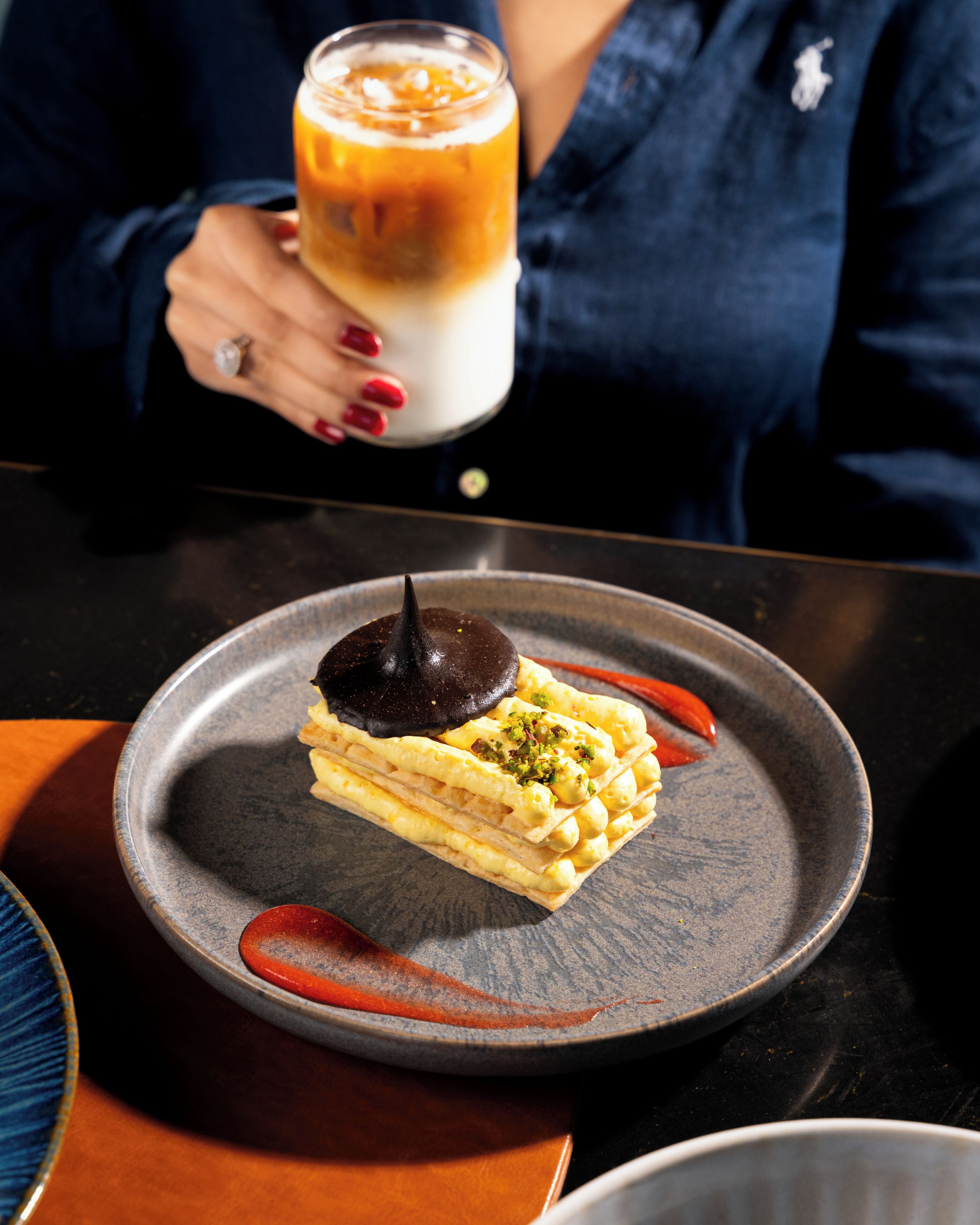 A dessert on a plate with a dark chocolate piece on top, a glass of a layered beverage, and a person with red nail polish.