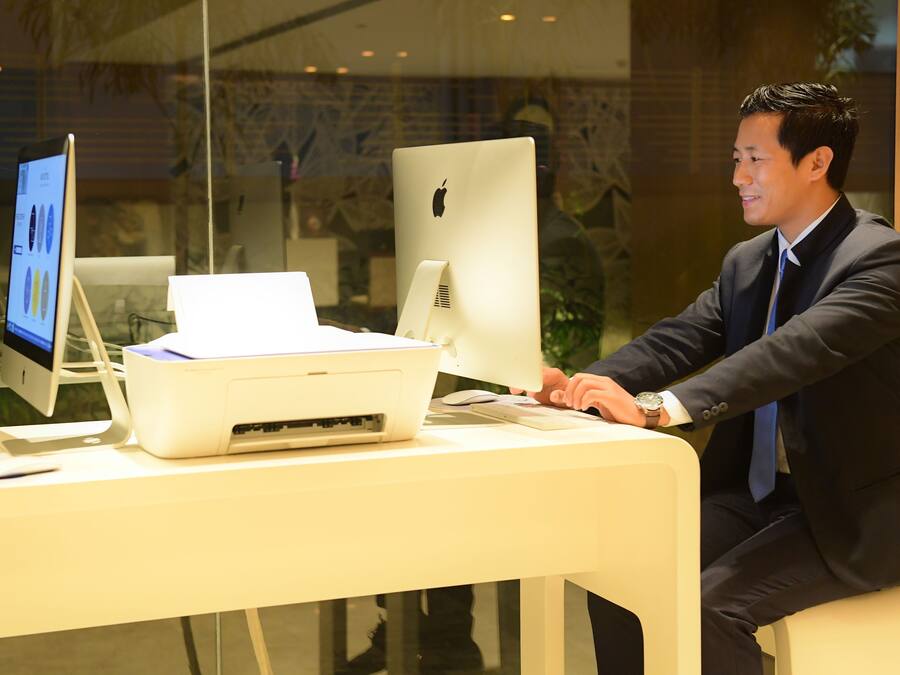 A man in a suit using a desktop computer with a monitor, keyboard, and printer on a desk.