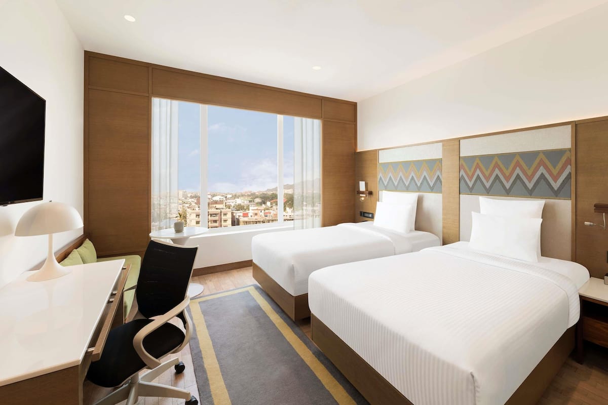 A hotel room with two beds, a desk, a chair, a TV, and a large window offering a city view.