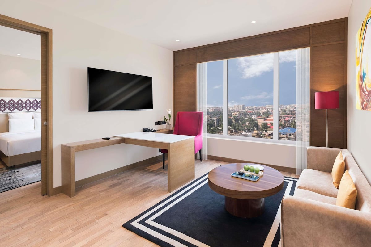 A modern hotel room with a bed, a desk, a TV, a sofa, a coffee table, and a large window with a city view.