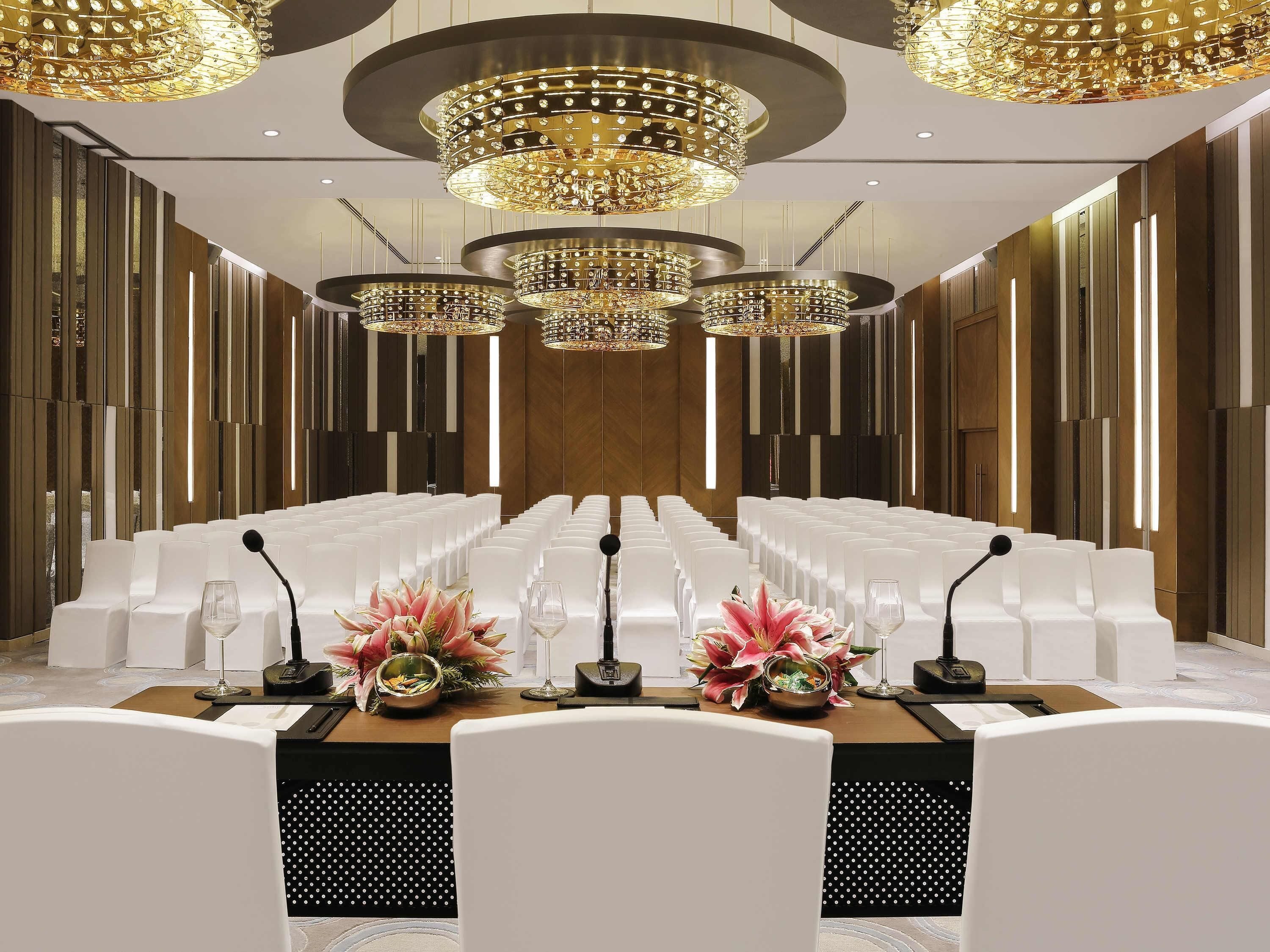 A conference room with rows of white chairs, a long table with microphones, and ornate chandeliers.