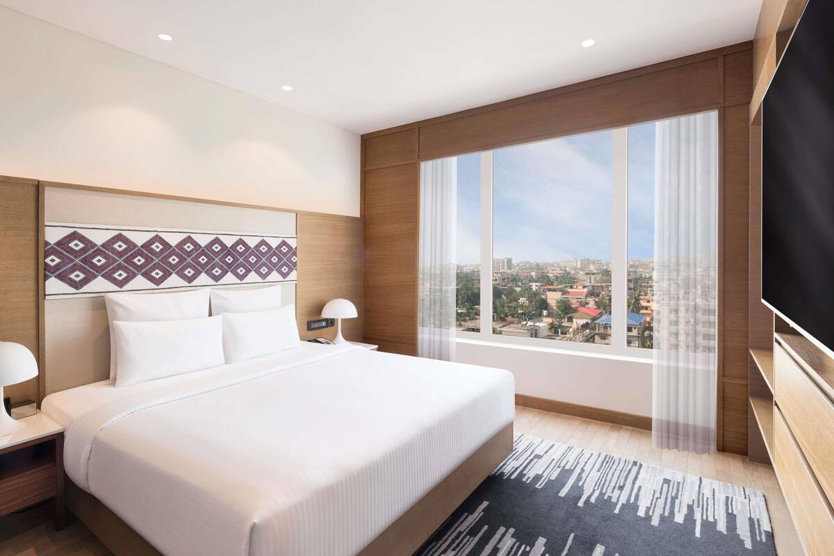 Una habitación de hotel con una cama grande, televisión, mesita de noche, lámpara, ventana con vista a la ciudad y cabecera estampada.