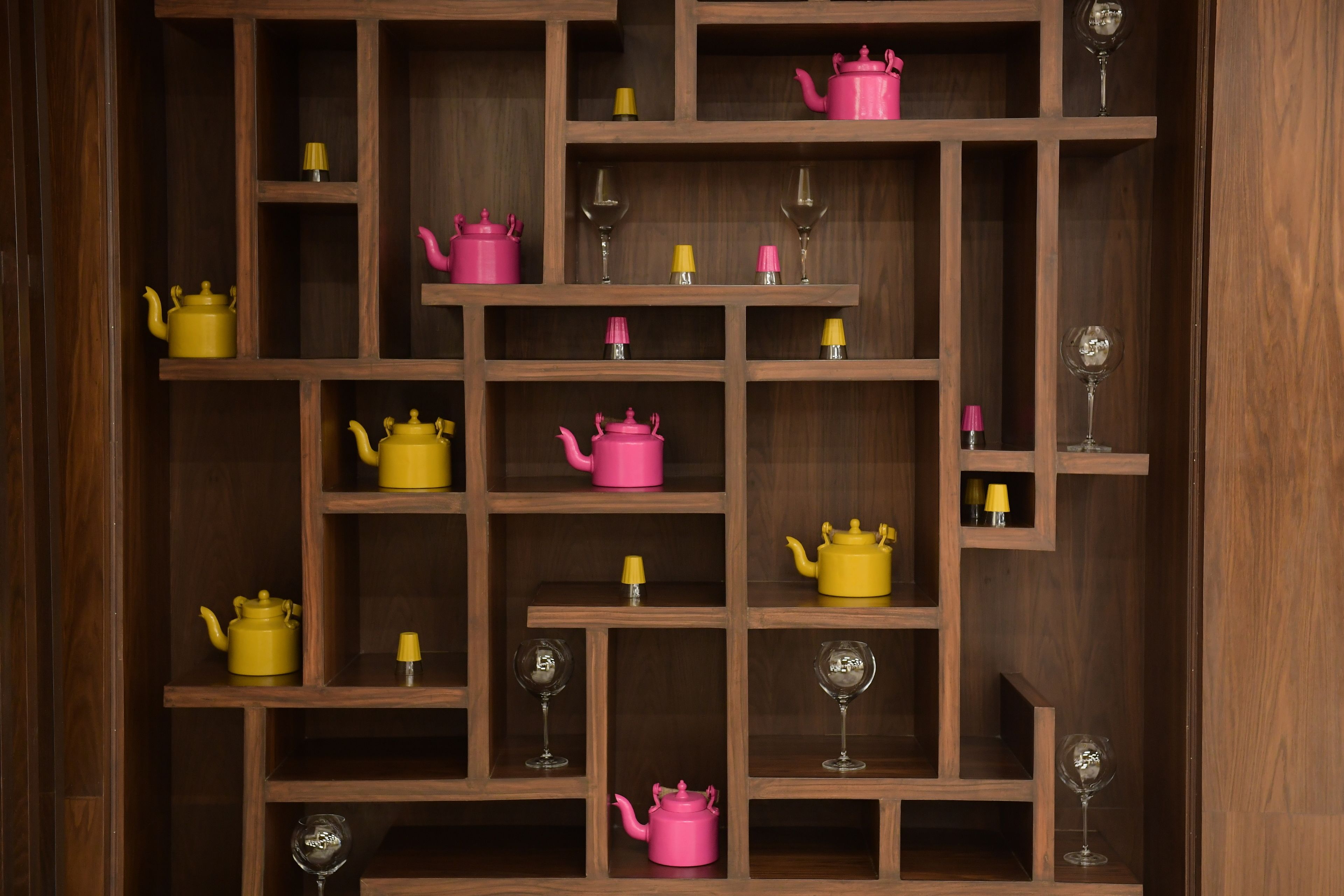 A wooden shelf unit with multiple compartments, each containing a pink teapot, a yellow teapot, and a wine glass.