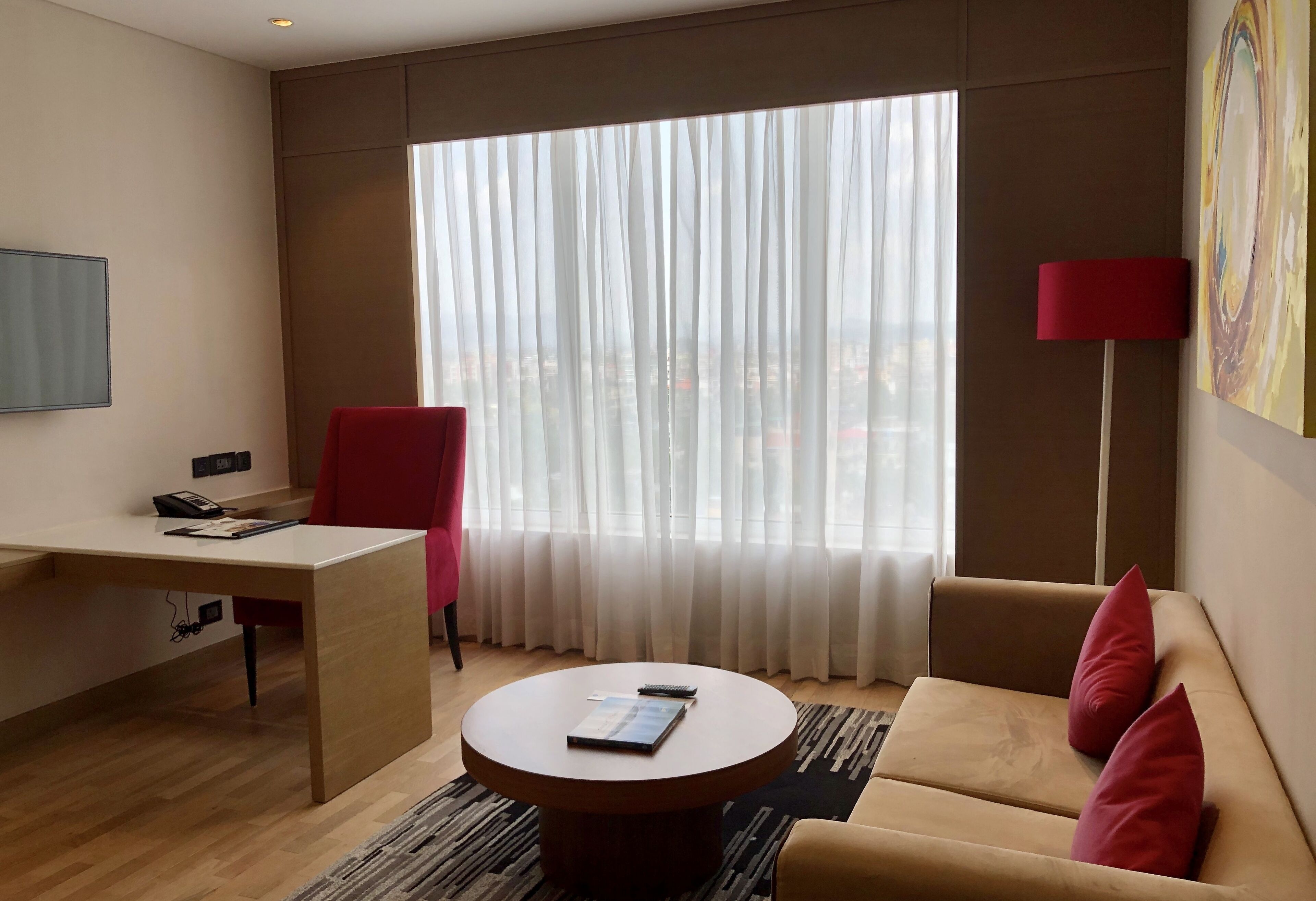 A hotel room with a wooden desk, a red chair, a round coffee table, a sofa with red cushions, a large window with sheer curtains, and a red floor lamp.