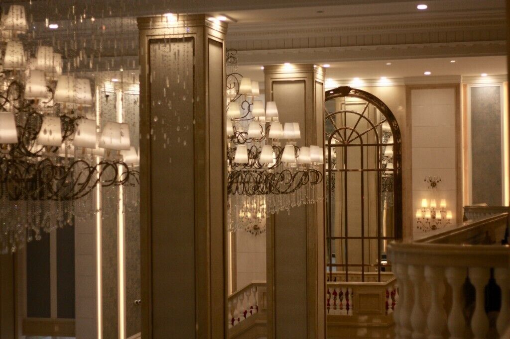 A grand lobby with ornate chandeliers, a large arched doorway, and a staircase with balustrades.