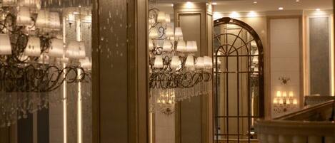 A grand lobby with ornate chandeliers, a large arched doorway, and a staircase with balustrades.
