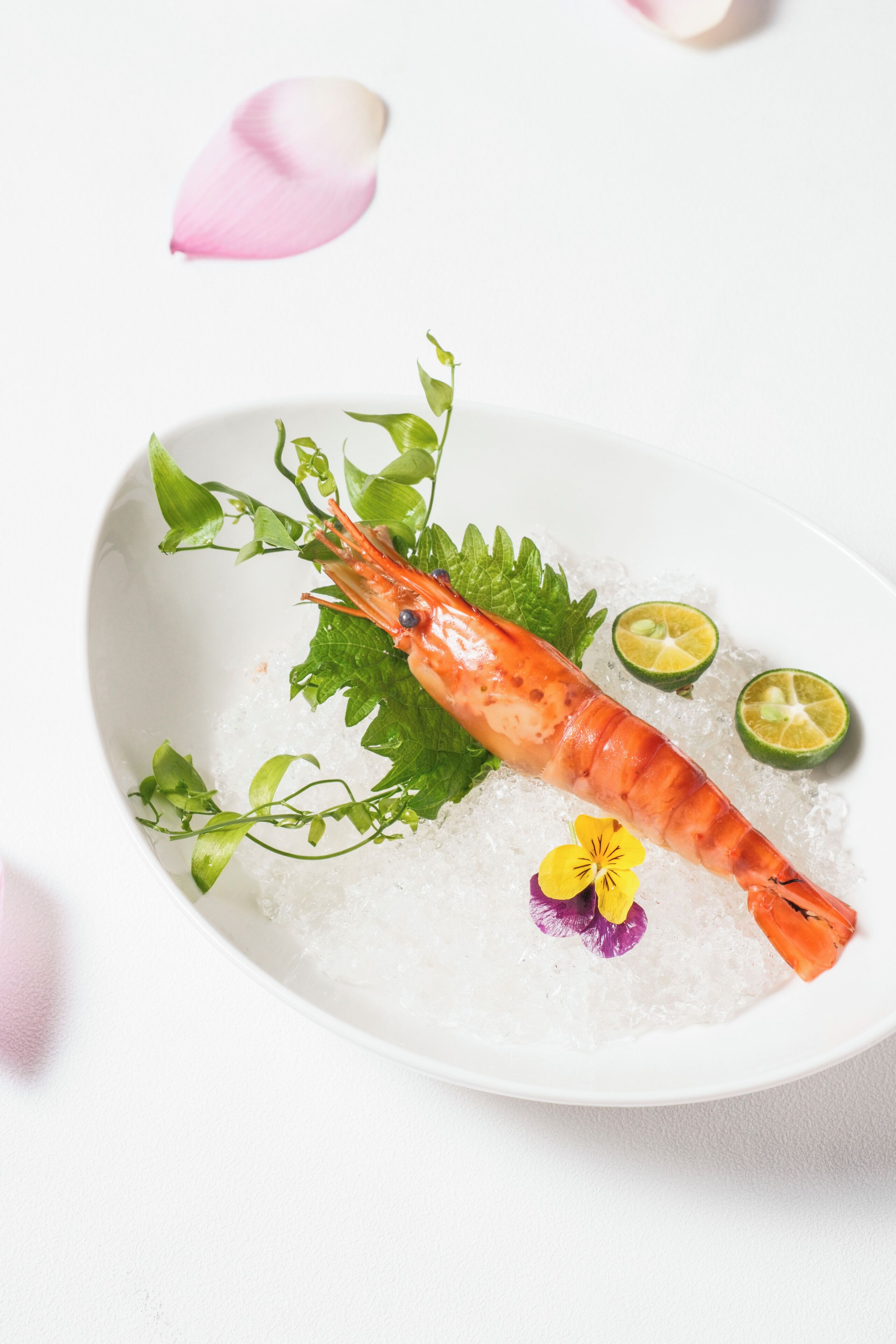 A bowl of ice with a cooked shrimp, lime slices, and decorative leaves.