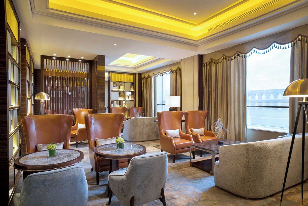 Un hall d’hôtel avec des fauteuils en cuir, une table ronde, une bibliothèque et une grande fenêtre donnant sur un beau paysage.